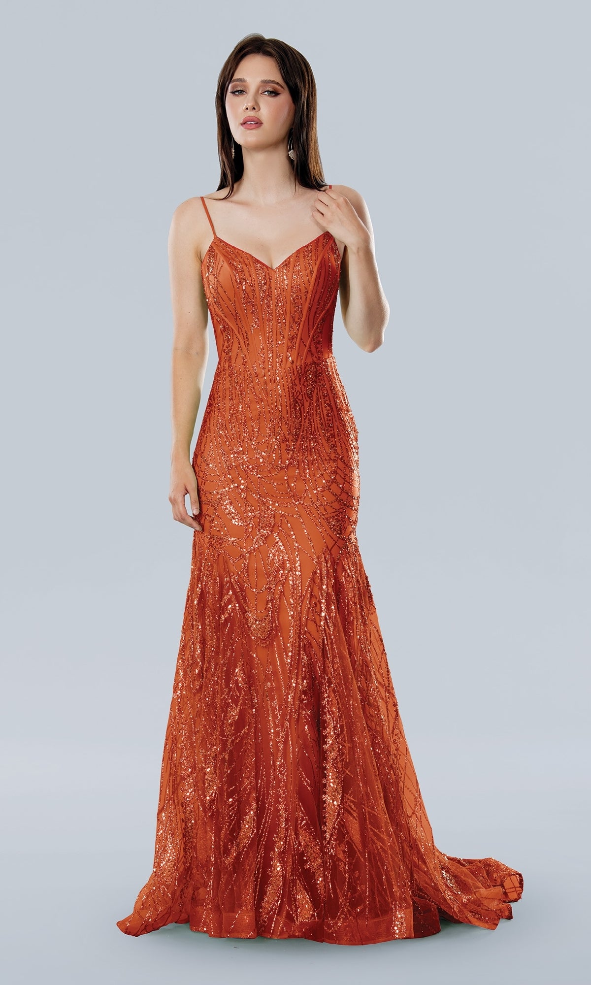 A woman models the Stella Couture 25105 long prom dress—an orange sequin gown with thin straps, a fitted floor-length silhouette, and a sheer back—standing against a plain light gray background.